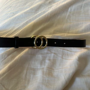 Svart bälte - Snyggt svart bälte med två runda guldfärgade ringar som spänne. Perfekt accessoar för att lyfta en outfit och ge en trendig touch. Bältet har klassisk design och flera hål för justerbar passform.