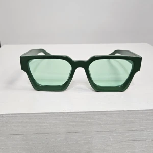 Gröna kantiga solglasögon - Snygga solglasögon med kraftig, mörkgrön båge och ljusgröna glas. De har en tydligt kantig och modern form som sticker ut. Perfekta för dig som vill ha en unik accessoar med attityd.