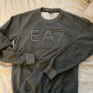 Grå sweatshirt från Emporio Armani - Säljer en stilren grå sweatshirt från Emporio Armani med broderad EA7-logga framtill. Tröjan har rund halsringning och långa ärmar med ribbade muddar. Perfekt för en avslappnad och snygg look.