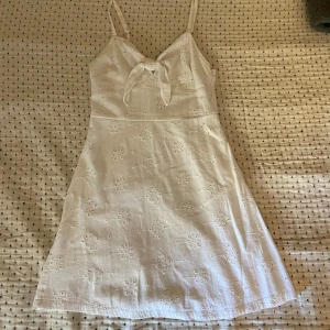 Vit sommarklänning - Söt vit klänning från Hollister med smala axelband och knytdetalj framtill. Klänningen har broderade blommor och är tillverkad i bomull. Klänningen har använts en del men är fortfarande i mycket bra skick.
