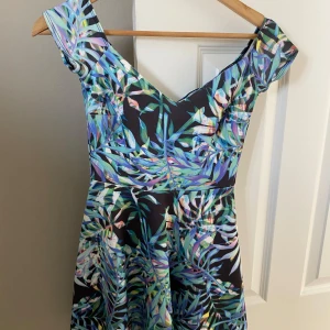 Blommig kortklänning från NLY One/Nelly - Säljer en färgglad kortklänning från NLY One med tropiskt blommigt mönster i blått, grönt och lila. Klänningen har offshoulder-modell och öppen rygg, perfekt för sommarens tillfällen. Materialet känns lätt och luftigt.