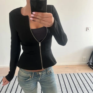 Snygg svart ”jacka” - Säljer en svart ”jacka” då jag ej andvänder den! Den har volang i botten och ger en snatched look. ”Jackan” har lång ärm och en silverfärgad dragkedja framtill. Enkel och stilren design som passar till både fint och vardag!
