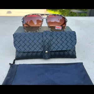 Bruna solglasögon med rutigt mönster - Snygga solglasögon med bruna tonade glas och rutigt mönster på bågarna. Metallbågar med UV 400-skydd. Kommer med ett svart, flätat fodral och en mjuk förvaringspåse. Perfekta för soliga dagar.HELT NYA - box och fodral ingår