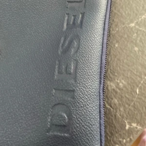 Mörkblå axelväska från Diesel - Snygg mörkblå axelväska från Diesel med präglad logotyp och motiv på framsidan. Väskan har justerbar axelrem, dragkedja och tryckknapp. Perfekt för att bära det viktigaste i vardagen.
