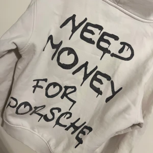 Vit hoodie med svart text - Vit hoodie med stor svart text på ryggen där det står 'Need money for Porsche' i graffiti-liknande stil. Tröjan har huva och ribbade muddar vid ärmslut och nederkant. Perfekt statement-plagg för dig som gillar streetwear.
