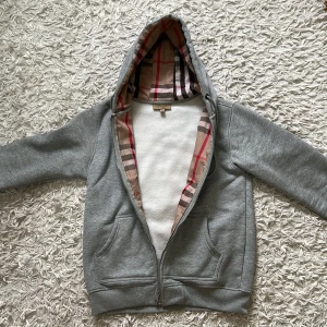 Burberry Zip up hoodie Grå - Storlek M, Passar perfekt, helt ny, och helt oanvänd, perfekt för våren och hösten, passar med allt. Embroiderad burberry logga på vänstra sidan, custom dragkedja med engraverad burberry logga, insidan och luva rutig- Se bild 1. Säljer pga för liten.