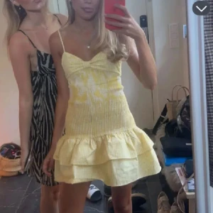 Gul volangklänning med smock och axelband - Söt gul klänning med smockad överdel och volangkjol. Axelbandsmodell med lätt rynk vid bysten och volanger nertill. Perfekt för varma dagar och somriga tillfällen.