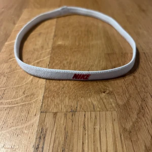 Vitt Nike diadem med röd logga - Snyggt vitt diadem från Nike med röd logga framtill. Smal och elastisk modell som håller håret på plats under träning eller vardag. Tillverkat i ett mjukt syntetmaterial som sitter bekvämt utan att glida. Perfekt accessoar för en sportig look.