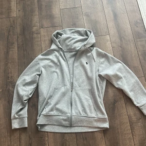 Grå hoodie från Polo Ralph Lauren - Snygg ljusgrå hoodie från Polo Ralph Lauren med dragkedja och klassisk broderad logga på bröstet. Tröjan har huva med snörning, två fickor fram och är tillverkad i mjuk bomullsmix. Perfekt för chill dagar eller när du vill ha en clean look.