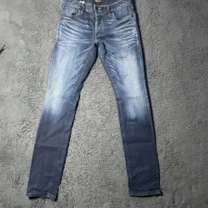 Slim fit jeans blå från Jack & Jones - Snygga slim fit jeans från Jack & Jones i mörkblå tvätt med slitningar och ljusare partier. Modellen heter Tim och har klassisk femficksdesign, knappgylf och smal passform. Perfekta för dig som gillar en modern och stilren look.