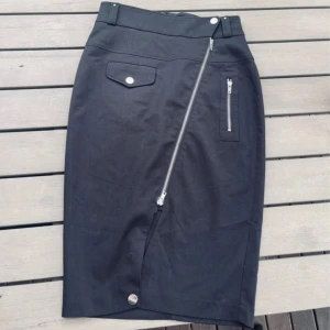 Svart kjol Jean Paul Gaultier x Lindex - Svart knälång kjol från Jean Paul Gaultier x Lindex med cool sned dragkedja framtill, fickdetaljer med silverknappar och bälteshällor. Kjolen har en stilren och edgy look, perfekt för dig som gillar statement-plagg.