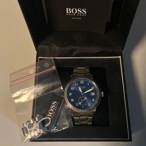 Blå och silverfärgad klocka från Hugo Boss - Stilren klocka från Hugo Boss med blå urtavla, datumvisning och silvrigt armband i metall. Klassisk rund form och tydliga siffror på urtavlan. Levereras i originalask med extra länk. Perfekt accessoar för dig som gillar snygga detaljer.