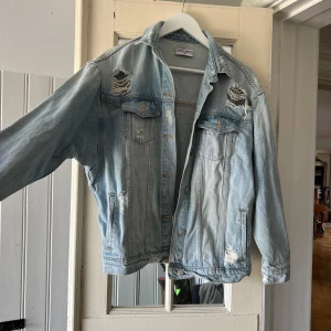 Ljusblå oversized jeansjacka - Säljer en ljusblå jeansjacka med slitna detaljer och klassisk krage. Jackan har två bröstfickor med lock, knappar framtill och en loose, oversized passform. Perfekt för dig som gillar en avslappnad streetstyle-look.