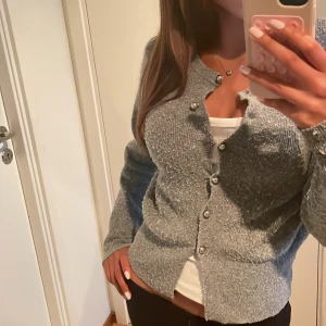 Grå stickad kofta med pärlknappar - Superfin grå stickad kofta med glansiga pärlknappar framtill. Koftan har långärmad passform och är lite fluffig i materialet, vilket ger en mysig och trendig look. Perfekt att slänga över ett linne eller t-shirt för en chill vibe.