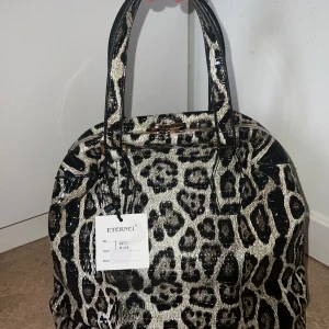 Leopardmönstrad handväska från Eternel - Snygg handväska från Eternel med glansigt leopardmönster i svart och beige. Väskan har två rejäla handtag, guldfärgade metalldetaljer och dragkedja upptill. Insidan är fodrad i satinliknande tyg med innerfack och dragkedja. Perfekt för dig som vill sticka ut.