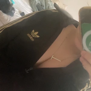 Svart Adidas track jacket med guld - Svart Adidas track jacket med guldiga ränder längs ärmarna och guldigt Adidas-tryck på bröstet. Klassisk sportig look med dragkedja framtill och långärmad passform. Perfekt för dig som gillar streetstyle och sportmode.