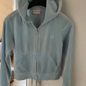 Ljusblå velour hoodie Juicy Couture - Supermjuk ljusblå hoodie från Juicy Couture i velour med dragkedja framtill, huva och snörning med gulddetaljer. Klassisk broderad JC-logga på bröstet och kängurufickor. Perfekt för en chill och trendig look.