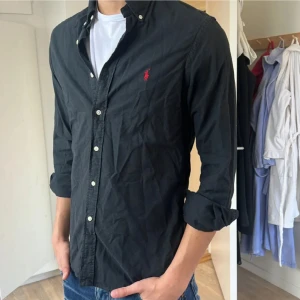 Ralph Lauren Skjorta (Pris diskuterbart) - !!NY SKICK!! Svart skjorta från Ralph Lauren i SLIMFIT  med klassisk passform och knappar framtill. Skjortan har lång ärm och ikonisk röd logga broderad på bröstet. Tillverkad i bomull och har button-down krage. Perfekt till jeans för en clean look.