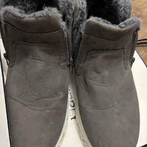 Grå boots med foder och dragkedja - Mysiga grå boots med mjukt foder och dragkedja på sidan. Skorna har en rund tå och vit sula, perfekta för kyliga dagar. Utsidan är i mockaliknande material och insidan är fodrad med fluffigt syntetpäls för extra värme.