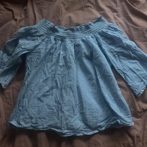 Blå offshoulder blus Twist & Tango - Säljer en ljusblå offshoulder blus från Twist & Tango i storlek 36. Blusen har tunna vita ränder, smockad kant upptill och trekvartsärmar. Perfekt för dig som gillar en luftig och avslappnad stil med lite boho-vibe.