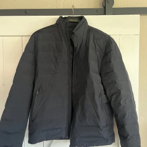 Svart pufferjacka med dragkedja - Svart pufferjacka från Mango med hög krage och två sidofickor med dragkedja. Jackan har quiltad design och stängs med hel dragkedja framtill. Perfekt för kalla dagar och enkel att matcha med olika outfits.