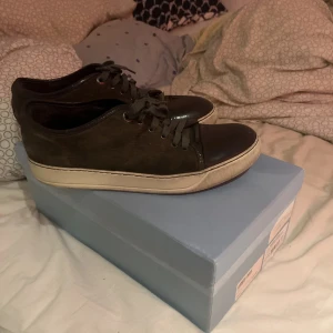 Bruna sneakers från Lanvin i mocka - Snygga bruna sneakers från Lanvin med ovandel i mocka och tåparti i blankt skinn. Skorna har rund tå, snörning och en beige platt sula som ger en clean look. Perfekta för dig som gillar stilrena och exklusiva sneakers.