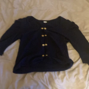 Mörkblå kofta från Bik Bok med knappar - Mörkblå kofta från Bik Bok i stickat material med snygga guldiga knappar framtill. Modellen har trekvartsärm och lätt puff vid axlarna för en trendig look. Perfekt att slänga över en topp när det blir kyligt. Säljes i storlek M.
