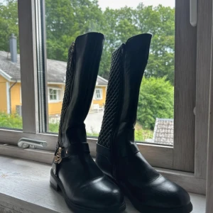 Svarta stövlar med dragkedja - Snygga svarta stövlar med   guldfärgade detaljer. Stövlarna har rund tå, platt sula och dragkedja längs sidan. Dekorativt spänne i guld vid ankeln ger en extra touch. Perfekta för dig som gillar stilrena boots med lite edge.