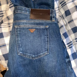 Emporio Armani blå jeans - Snygga blå jeans från Emporio Armani med klassisk femficksdesign och ikoniska logodetaljer i metall på bakfickan och myntfickan. Jeansen har en normal passform och är tillverkade i bomull med knappgylf. Perfekta för dig som gillar stilrena och exklusiva jeans.