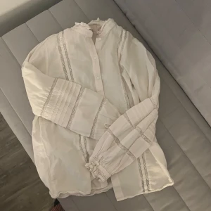 Vit romantisk blus med spetsdetaljer - Supersöt vit blus med spetsdetaljer och knappar framtill. Långärmad modell med rynkade ärmslut och dekorativa band längs ärmar och framstycke. Perfekt för dig som gillar en romantisk och vintage-inspirerad stil.