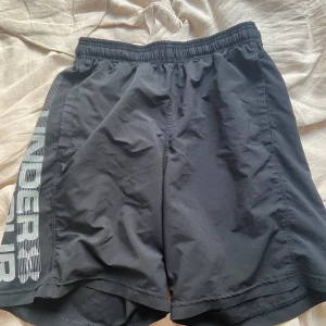 Svarta träningsshorts Under Armour - Svarta träningsshorts från Under Armour med stor logga längs ena benet. De har elastisk midja med snörning och är tillverkade i ett lätt och snabbtorkande syntetmaterial. Passformen är loose och perfekta för gymmet eller löprundan.