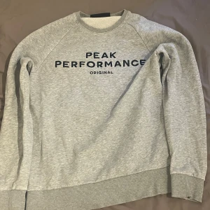 Grå sweatshirt från Peak Performance - Säljer en stilren grå sweatshirt från Peak Performance med svart logga framtill. Tröjan har rund hals, ribbade muddar och är tillverkad i mjuk bomullsmix. Perfekt för en chill och sportig look.