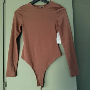 Brun långärmad body  - Säljer en stilren brun långärmad body från Biancas gamla kollektion med Nelly. Modellen har rund halsringning och är figurnära med klassisk bodyform. Har tyvärr aldrig kommit till användning så prislappen sitter kvar.