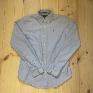 Ralph Lauren Skjorta  - Klassisk ljusblå skjorta från Ralph Lauren i slim fit-modell. Skjortan har button down-krage, långa ärmar och broderad polospelare på bröstet. Tillverkad i 100% bomull och stängs med vita knappar framtill. Perfekt för en clean och stilren look.