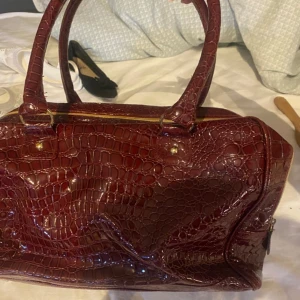 Vinröd handväska i crocodil - Säljer en mörkröd handväska med croco-mönstrad yta och guldiga detaljer. Väskan har två stadiga handtag, dragkedja upptill och ett rymligt innerfack. Perfekt för dig som vill sticka ut med en elegant och trendig accessoar.