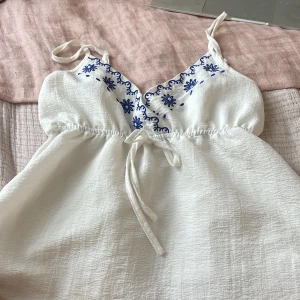 Vit broderad linnetopp från SHEIN - Superfin vit linnetopp från SHEIN med blå broderade blommor längs urringningen. Toppen har smala axelband, knytning under bysten och en lätt strukturerad yta. Perfekt för en fräsch och somrig look! Aldrig använd