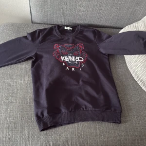 Svart Kenzo sweatshirt med tiger - Svart sweatshirt från Kenzo med ikoniskt broderat tigerhuvud och logga på bröstet. Tröjan har rund hals, långa ärmar och ribbade muddar. Perfekt statement-plagg för dig som gillar streetstyle och vill sticka ut.