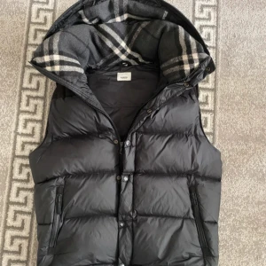 Svart dunväst från Burberry XL - Svart dunväst från Burberry med avtagbara ärmar och huva. Västen har dragkedja och knappar framtill, två fickor med dragkedja och klassiskt rutigt foder i huvan. Perfekt för kalla dagar och riktigt snygg streetstil. Kan mötas upp i Sthlm eller frakta om det behövs.