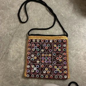 Broderad svart axelväska med pärlor - Unik svart axelväska med broderade färgglada pärlor och guldiga detaljer. Väskan har fyrkantig form, dragkedja upptill och flätad axelrem. Framsidan är täckt av blommönster i rött, vitt, lila, grönt och gult. Perfekt för dig som vill sticka ut med accessoarer.