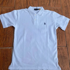 Ralph Lauren pike - Klassisk vit pikétröja från Ralph Lauren med broderad marinblå logga på bröstet. Tröjan har korta ärmar, krage och knäppning med två knappar. Tillverkad i mjuk bomull som känns skön mot huden. Perfekt för en clean och stilren look.