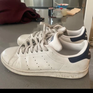 Adidas Stan Smith vita sneakers - Klassiska Adidas Stan Smith sneakers i vitt skinn med marinblå detalj på hälen och perforerade ränder på sidorna. Skorna har rund tå, platt sula och snörning framtill. Perfekta för en clean och tidlös stil.