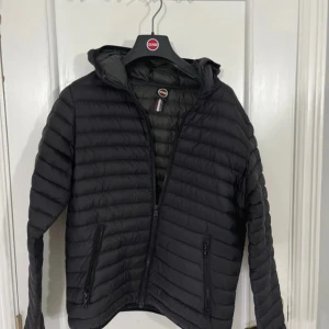 Svart dunjacka från Colmar - Snygg svart dunjacka från Colmar med horisontella sömmar och huva. Jackan har dragkedja framtill och två praktiska fickor med dragkedja. Perfekt för kyliga dagar och enkel att matcha med allt. Klassisk logga på ärmen ger en sportig vibe. Storlek 16 y eller xs 