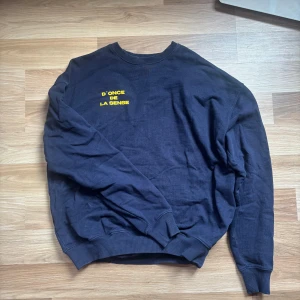 Mörkblå sweatshirt med gul text - Mörkblå sweatshirt med rund hals och ribbade muddar. På bröstet finns ett tryck i gult med texten 'D'ONCE DE LA SENSE' Alltså ”uns av förnuft” på svenska. Tröjan är långärmad och har en avslappnad passform, perfekt för en chill och stilren look.