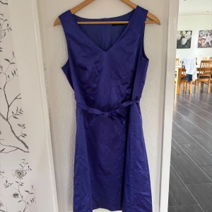 Blå/lila klänning från Esprit - Snygg blå,lila klänning från Esprit med v-ringning och knytband i midjan. Klänningen har en elegant, enkel design med dragkedja i ryggen. Bandet runt midjan går att ta väck. Använd några gånger men inga defekter. Ser blå ut i ljuset men har en blå lila nyans💜. Läs gärna bio😊