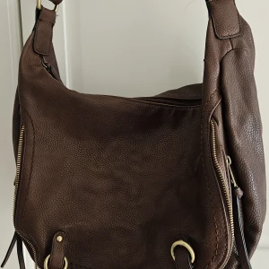 Brun axelväska från David Jones - Snygg brun axelväska i skinn från David Jones med guldfärgade metalldetaljer. Väskan har ett blommigt innerfoder och en rymlig design som gör den perfekt för vardagens alla prylar.