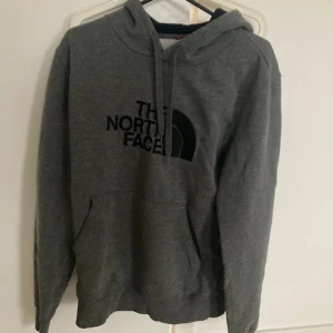 Grå hoodie från The North Face M - Säljer en grå hoodie från The North Face i storlek M. Tröjan har huva med snörning, stor magficka och svart logga framtill. Perfekt för chill dagar eller när du vill ha en enkel och stilren look. Mjuk och skön bomullsblandning.