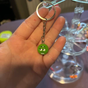 Grön M&M nyckelring - Söt nyckelring med en grön M&M-figur som hänge. Den lilla bollen har ett ansikte med busig blick och klassiskt vitt 'm' på framsidan. Perfekt accessoar för dig som gillar godis eller vill ha något kul på nyckelknippan.