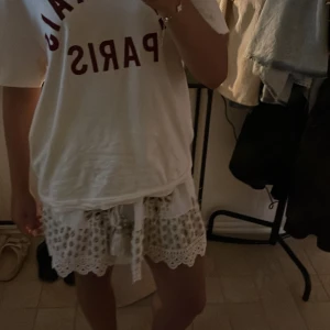 Vit kortkjol med broderi och snörning - Superfin vit kortkjol med bruna broderade detaljer och mönster, volangkant och snörning med tofsar och pärlor i midjan. Kjolen har en bohemisk vibe och är perfekt för varma dagar. Materialet känns lätt och luftigt, och spetsdetaljerna längst ner ger extra stil.