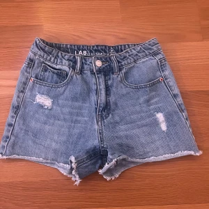 Blå jeansshorts från LAB Industries - Snygga blå jeansshorts från LAB Industries med hög midja, slitna detaljer och fransig kant nertill. Klassisk femficksmodell i denim med dragkedja och knapp. Perfekta för sommaren och ger en cool, avslappnad vibe.