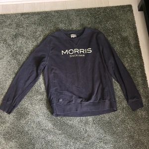 Mörkblå sweatshirt från Morris - Snygg mörkblå sweatshirt från Morris med vit logga framtill. Klassisk rund halsringning och långa ärmar. Perfekt för en chill och stilren look. Tillverkad i mjuk bomull som känns skön mot huden.